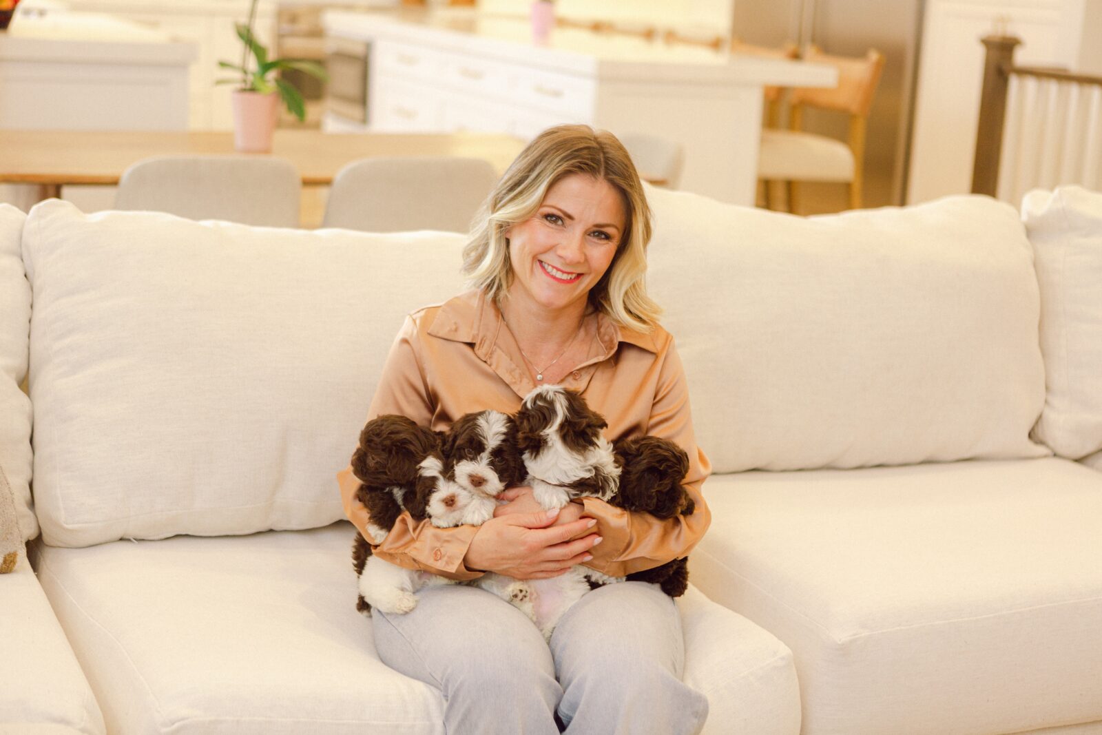 Cassie Milar smiling on a cream sofa while holding brown and white puppies.