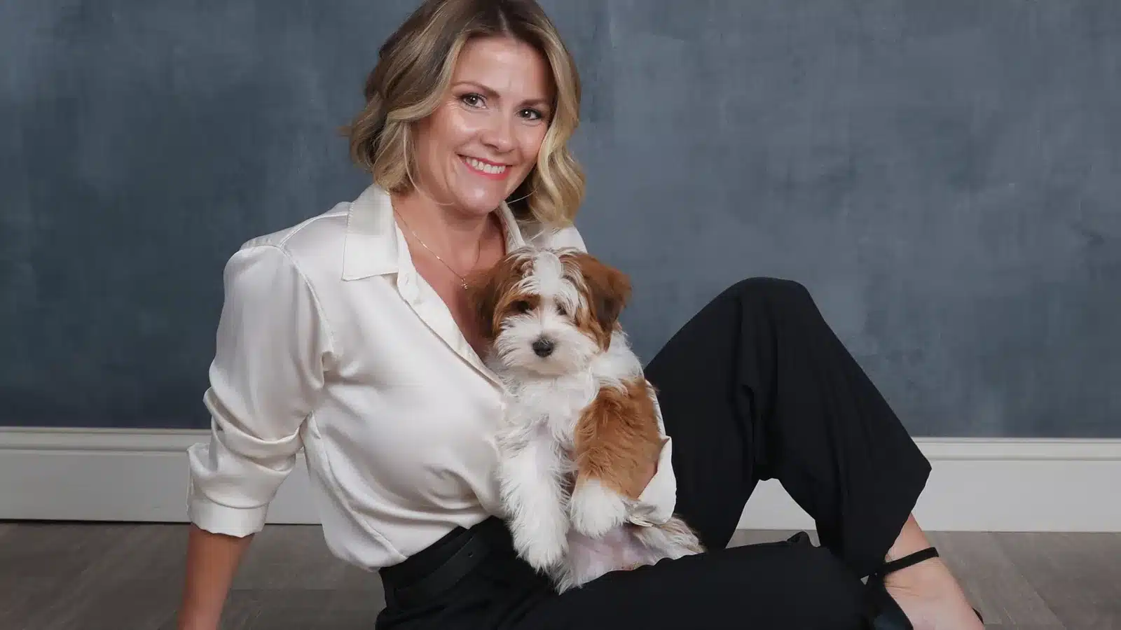 A smiling woman with blonde hair sits cross-legged on the floor, holding a small, fluffy brown and white puppy. She wears a silky white blouse and black pants, with a dark, textured backdrop.