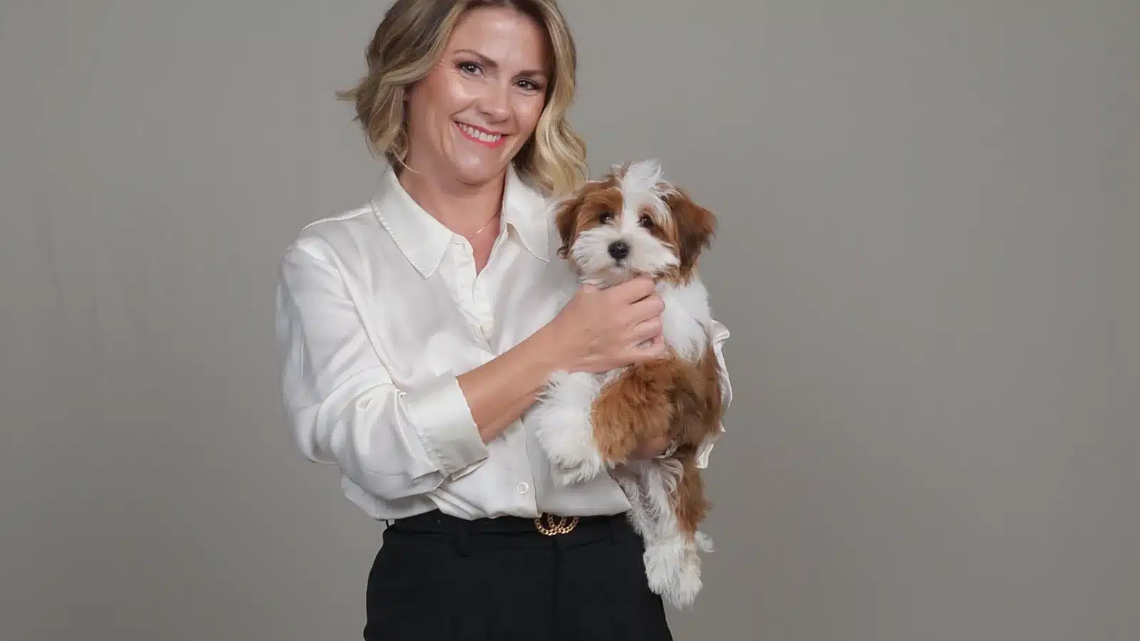 A smiling blonde woman in a white satin blouse and black pants holds a fluffy, brown and white Havanese puppy in front of a neutral gray background.